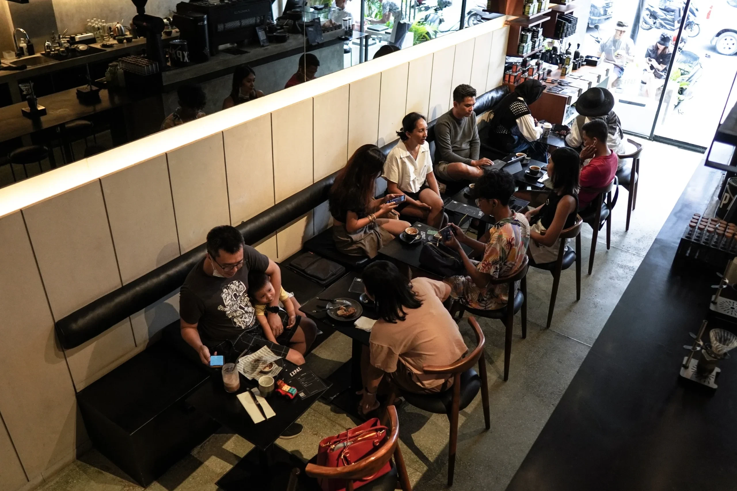 High-angle view of a busy modern cafe with customers sitting at small black tables on a long banquet bench, creating a vibrant community atmosphere.