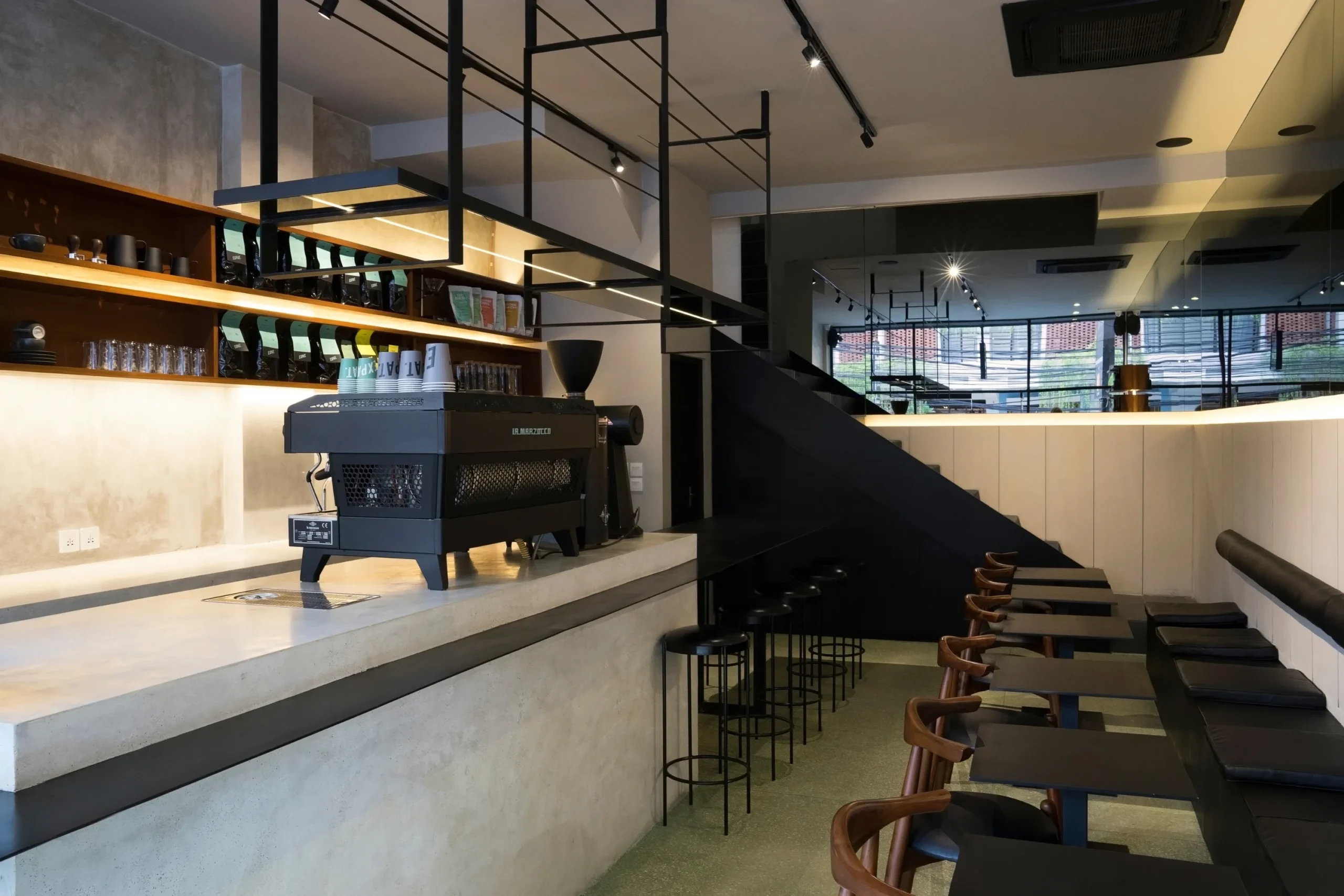 An industrial-style cafe interior featuring a concrete coffee bar, a professional black espresso machine, and a modern seating area with minimalist furniture and dark accents.