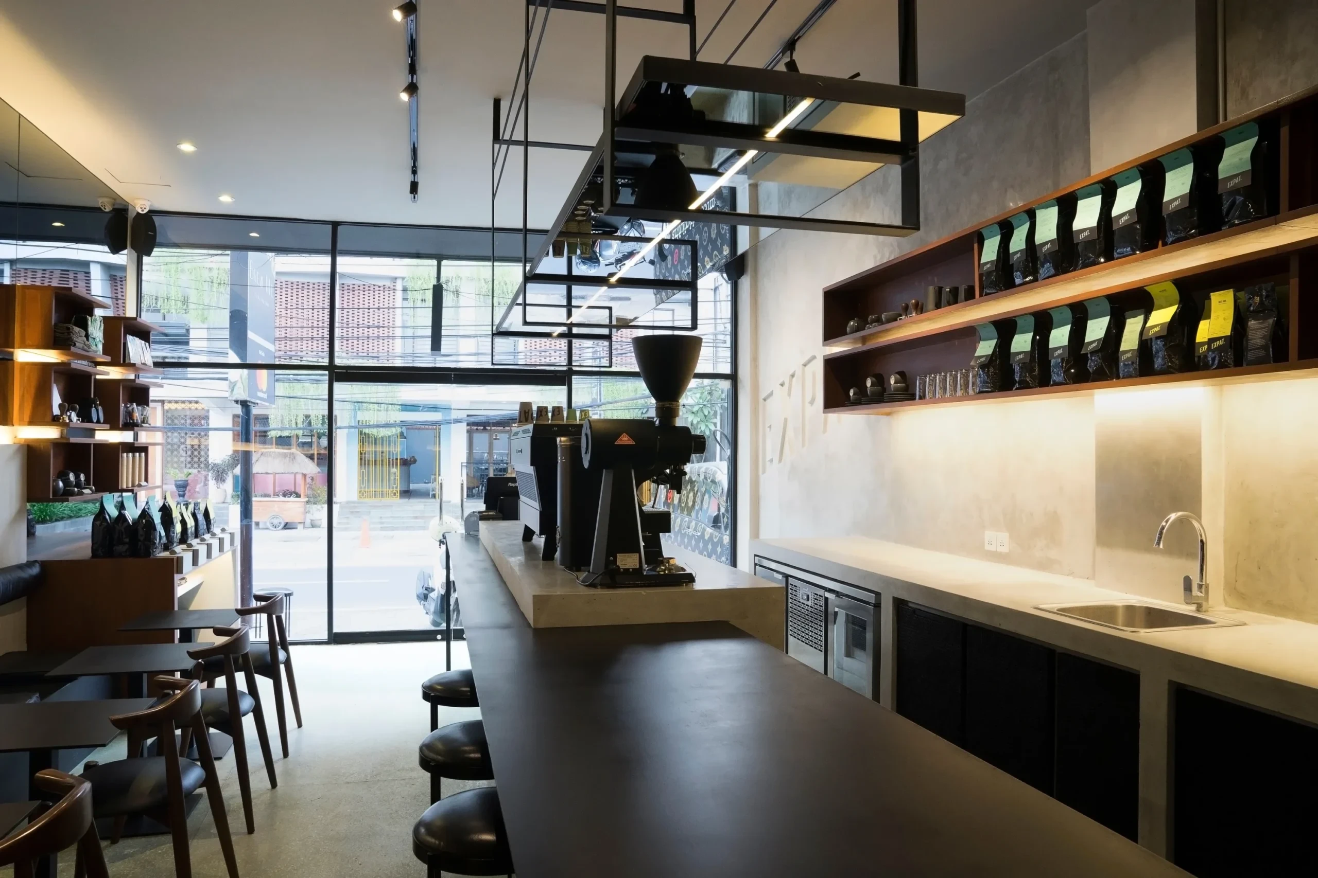 An industrial-style cafe interior featuring a concrete coffee bar, a professional black espresso machine, and a modern seating area with minimalist furniture and dark accents.