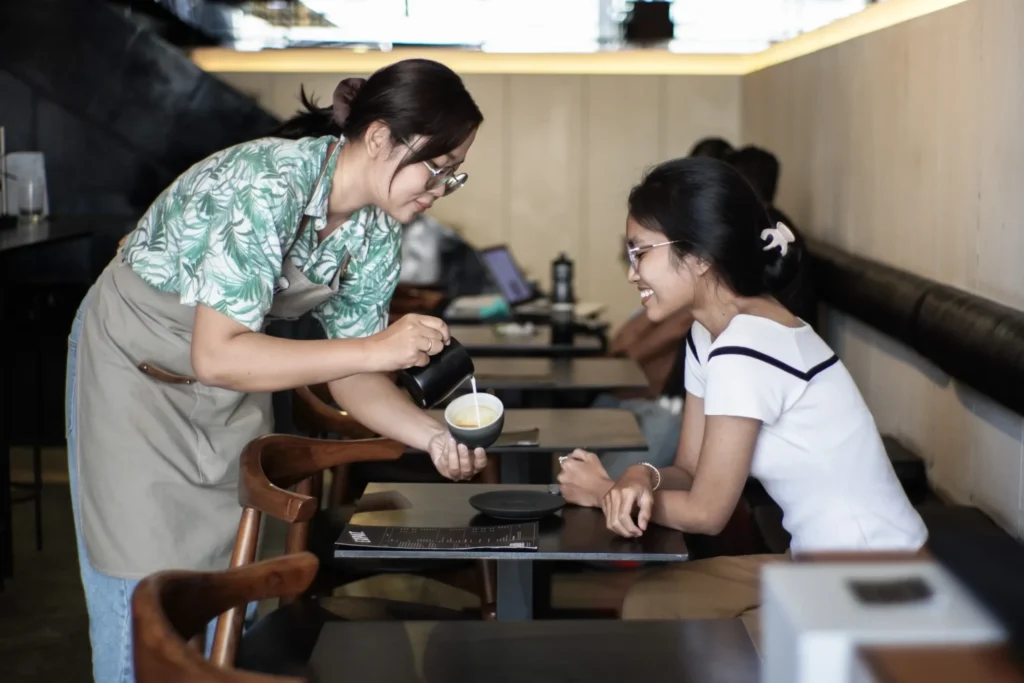 A barista in a tropical print shirt pouring latte art into a cup for a smiling female customer at a modern cafe table.