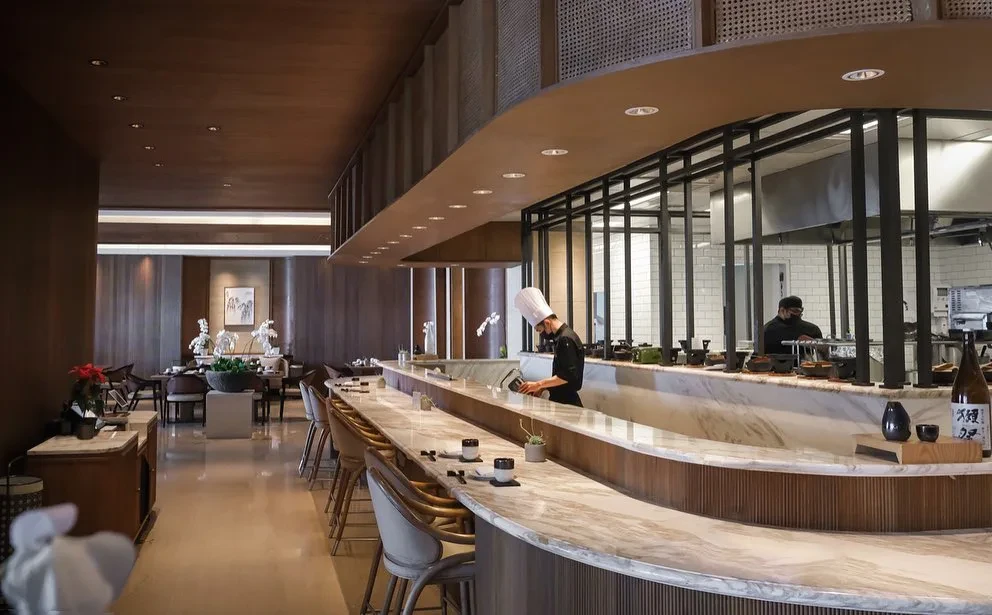 A modern Japanese restaurant interior featuring a long, curved marble sushi bar with seating and a chef working behind the counter.