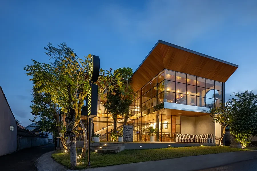 A modern, two-story glass-walled building at dusk with warm interior lighting, a prominent wooden cantilevered roof, and lush green landscaping.