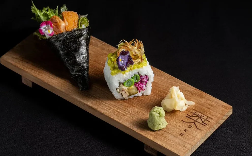 A wooden platter with a sushi hand roll, a maki roll, wasabi, and ginger.