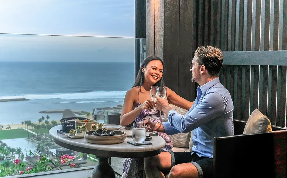 A man and woman toast with white wine while dining on an outdoor balcony overlooking a coastal beach and ocean at dusk.